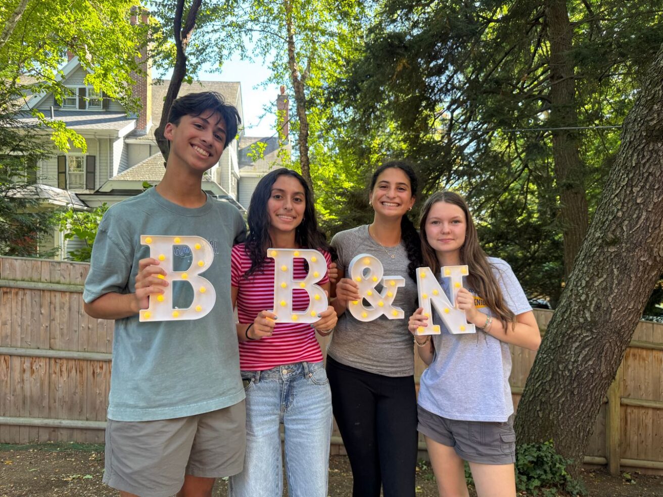 Four young people stand outdoors holding large illuminated letters that spell “B & N.” Trees and a wooden fence are visible in the background.