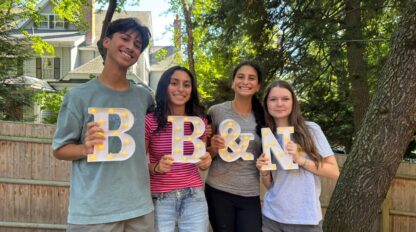 Four young people stand outdoors holding large illuminated letters that spell “B & N.” Trees and a wooden fence are visible in the background.