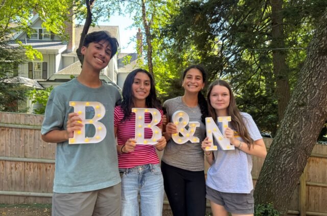 Four young people stand outdoors holding large illuminated letters that spell “B & N.” Trees and a wooden fence are visible in the background.