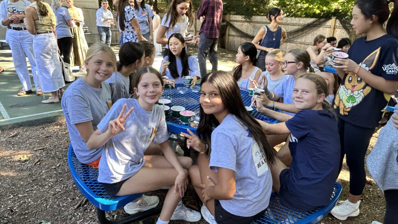 A group of girls sit and stand around a blue picnic table outdoors, eating and smiling. Other people gather in the background near a basketball court.