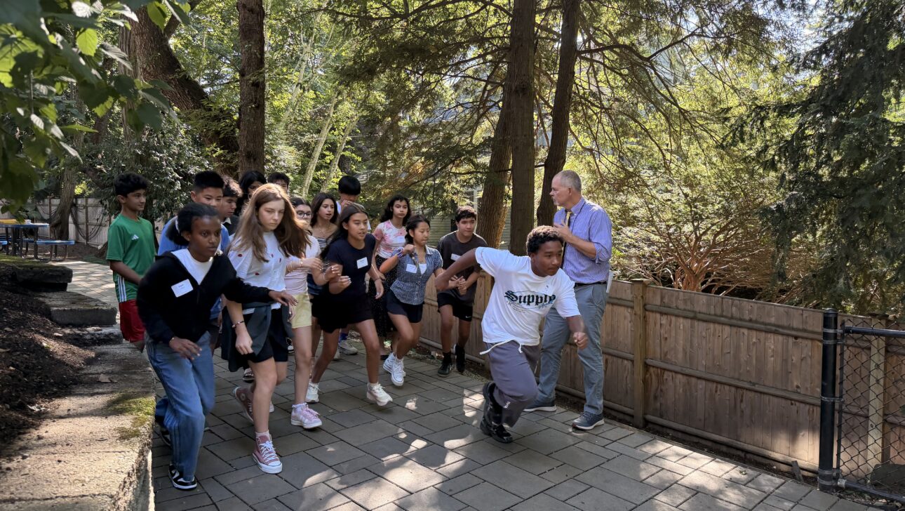 A group of students and one adult are running together on a paved walkway in a wooded outdoor area.
