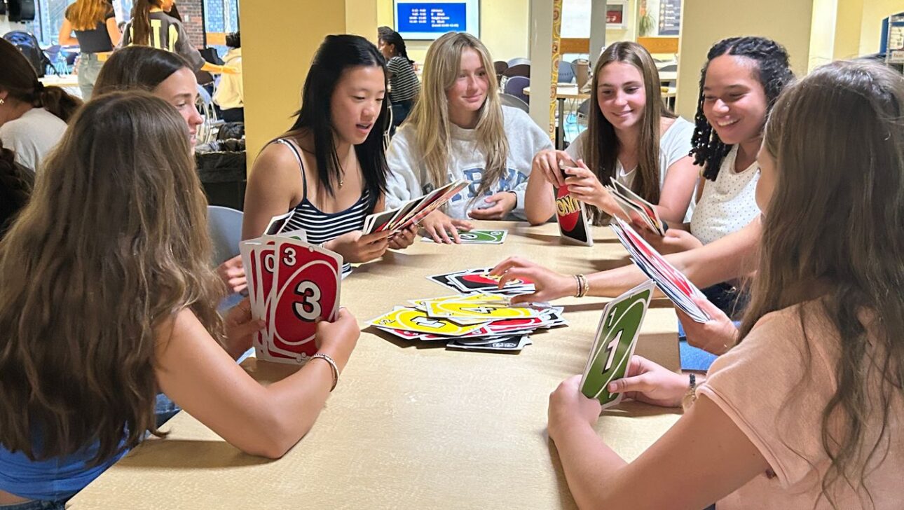 Seven young women sit around a table playing a card game, each holding large cards. The setting appears to be an indoor communal space.