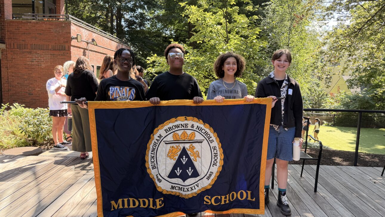 Four students stand outdoors holding a large “Middle School” banner, with two more people in the background. The setting is sunny with trees and a brick building visible.