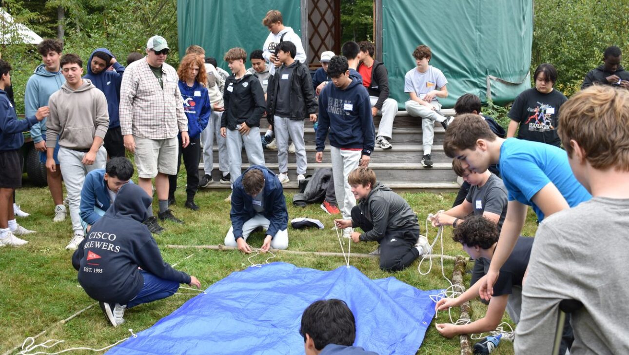 A group of teens and adults gather outdoors, some tying ropes to a blue tarp spread on the grass, with a wooden structure and trees in the background.