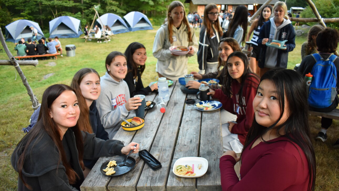 A group of teenage girls sits at a picnic table outdoors eating food, with tents and other people visible in the background at a campsite.