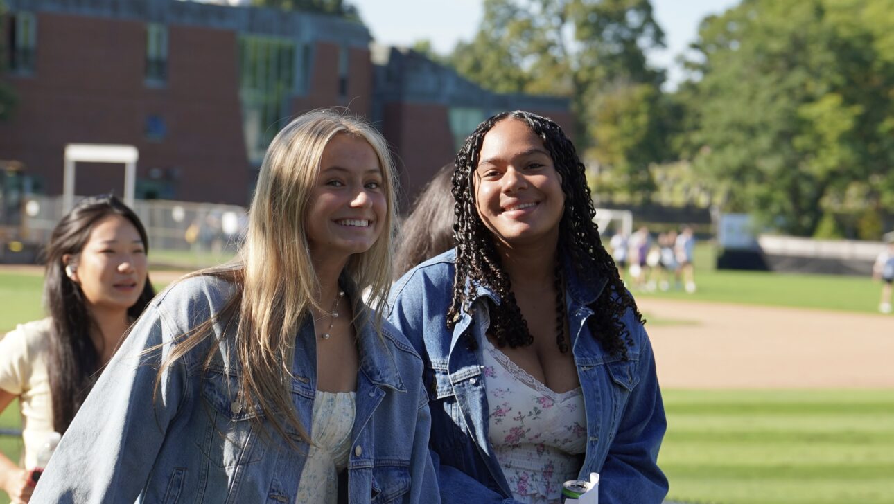 Two young women in denim jackets stand and smile outdoors near a sports field, with another person walking behind them and a brick building in the background.