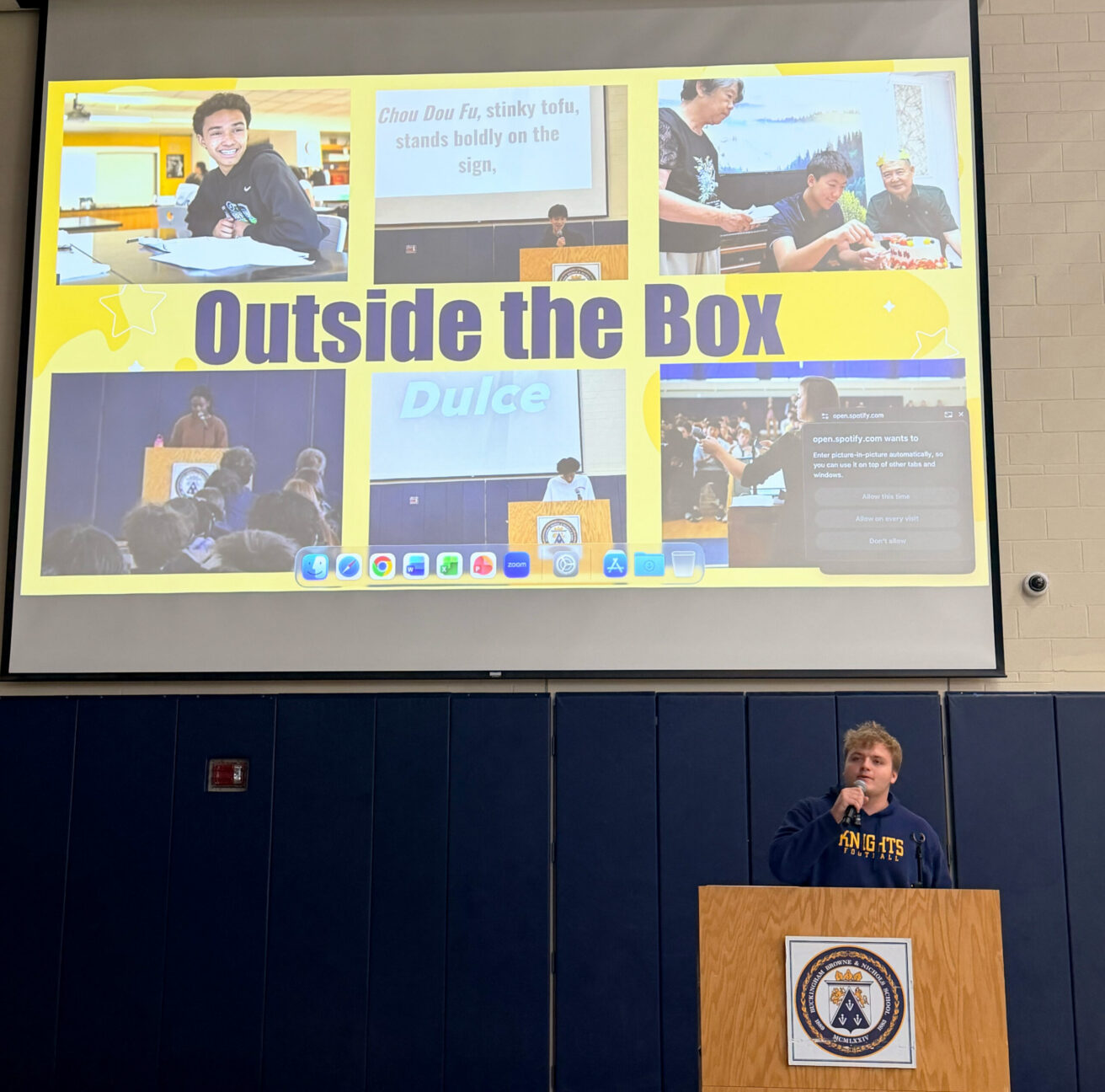 A person speaks at a podium in front of a large screen displaying a collage titled “Outside the Box” with photos and text highlights.