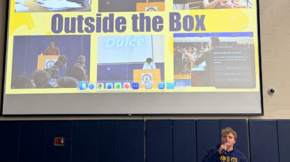 A person speaks at a podium in front of a large screen displaying a collage titled “Outside the Box” with photos and text highlights.