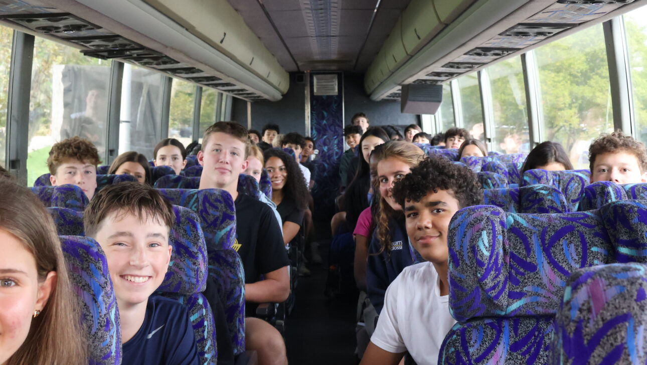 A group of teenagers sit on a coach bus facing forward, smiling and talking, with patterned seats and large windows showing greenery outside.