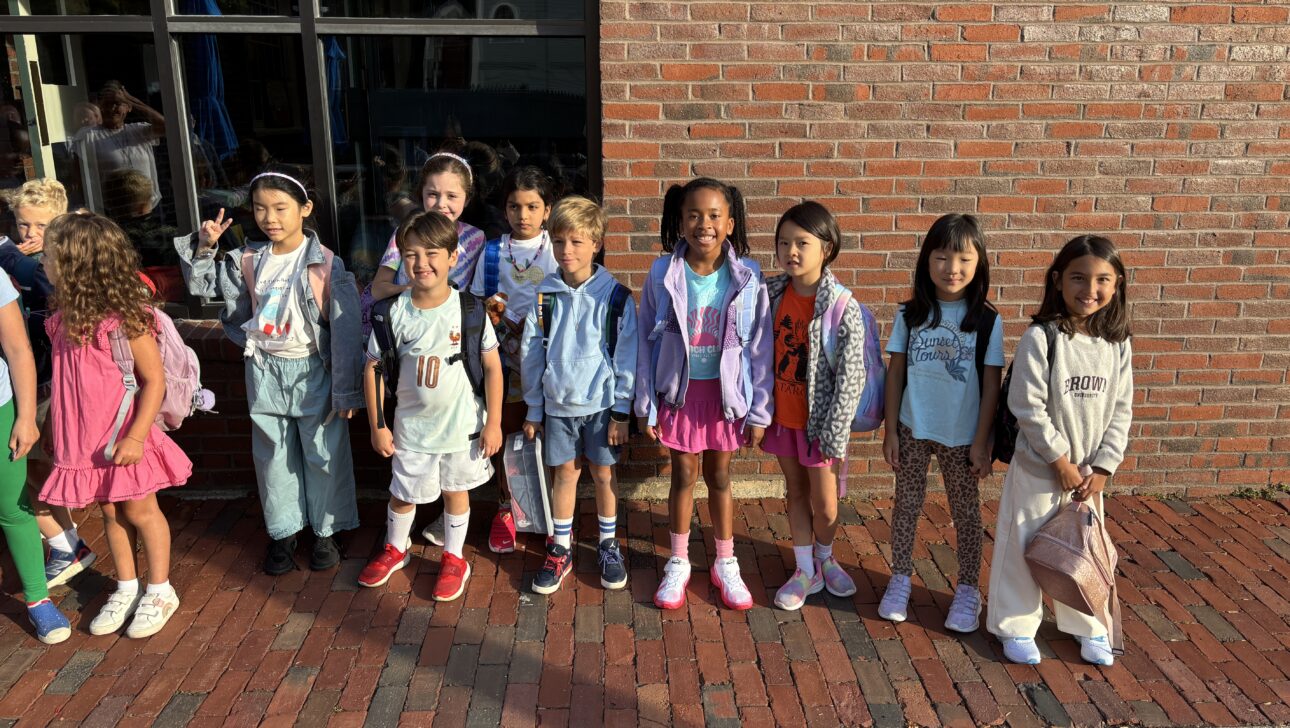 A group of eleven young children with backpacks stand in a row outside a brick building, facing the camera in morning sunlight.