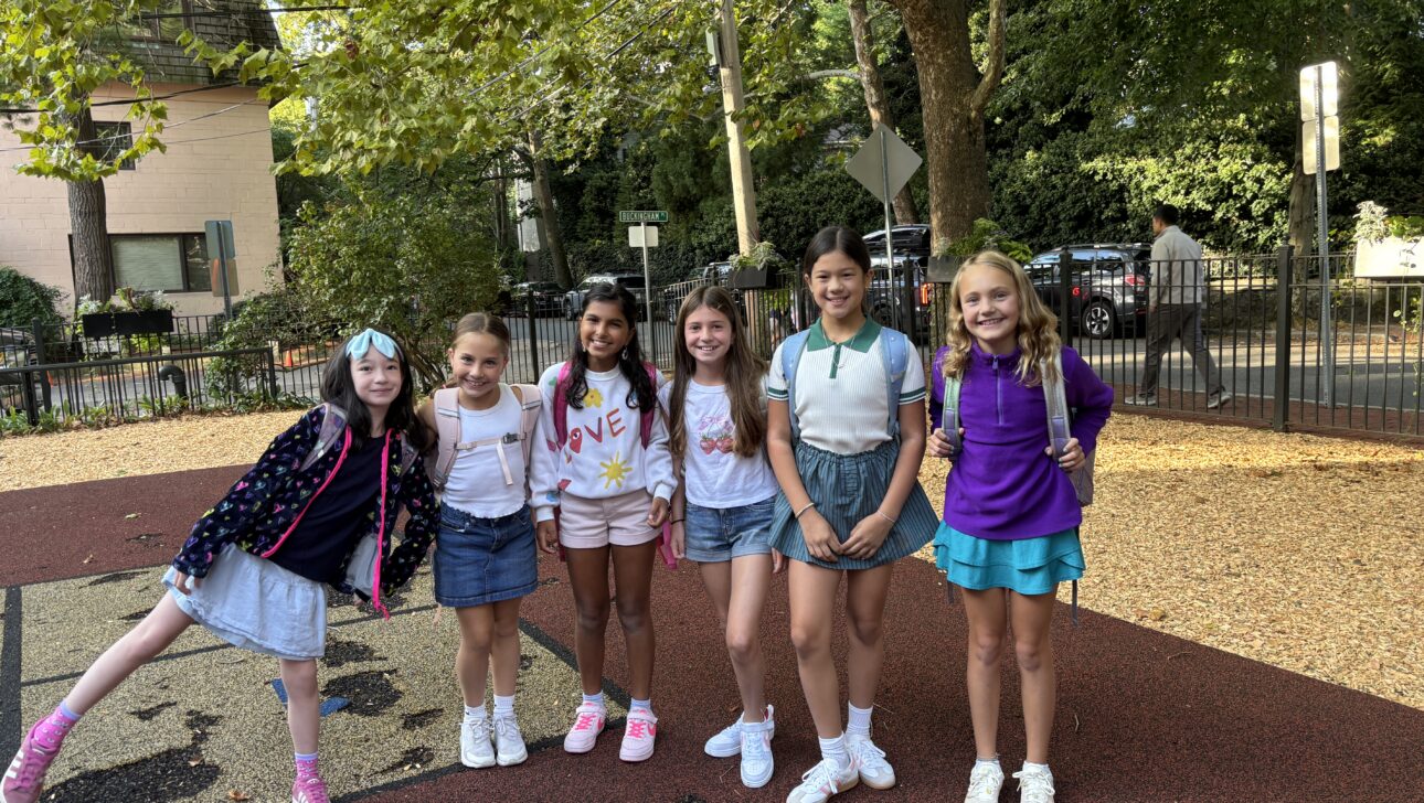Six girls stand in a row outdoors on a playground, smiling at the camera. Trees and parked cars are visible in the background on a sunny day.