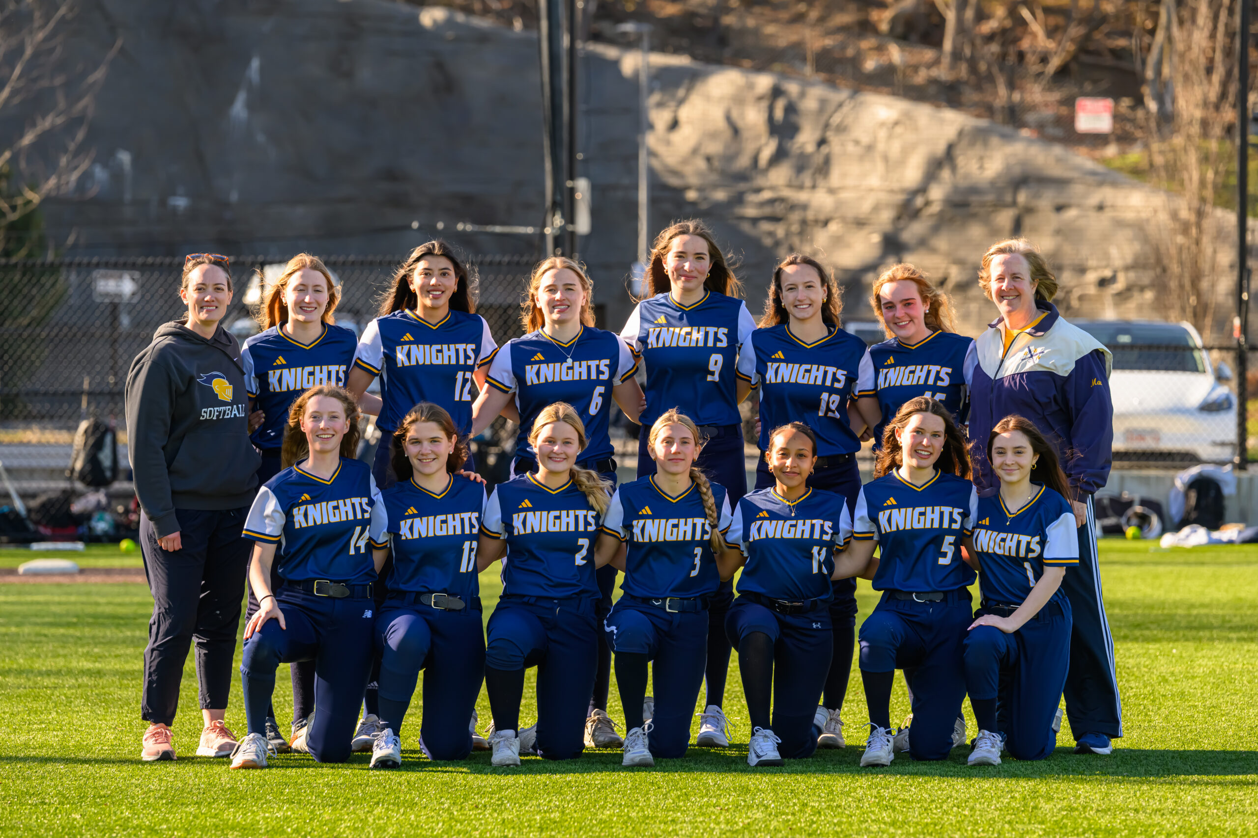 A girls’ softball team in blue “Knights” uniforms poses together on a sunny field, with two coaches standing at each end of the group.