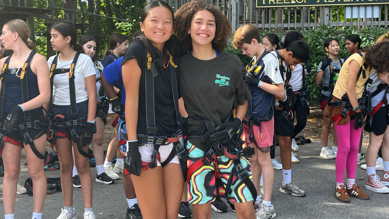 Two teens stand smiling in front of a group at an outdoor ropes course, wearing harnesses and gloves. A sign reading “TreeTop Adventure” is visible in the background.