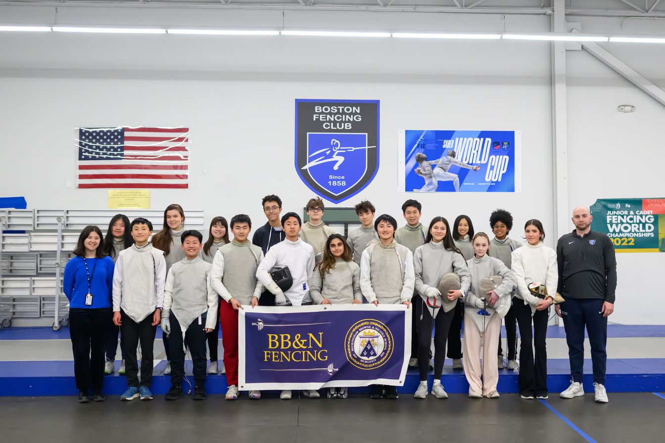 A group of young fencers and coaches pose indoors at a fencing club, holding a “BB&N Fencing” banner, with fencing gear and banners in the background.